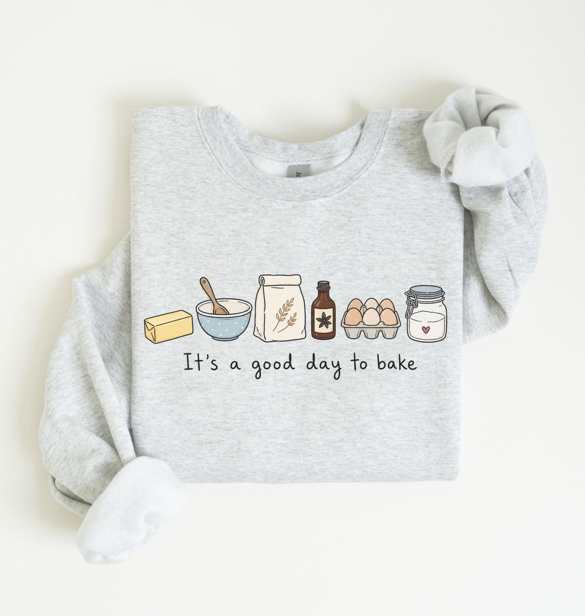 A light heather gray long-sleeve sweatshirt with a graphic of baking ingredients and the text 'It's a good day to bake' on the front, styled flat on a light cream background.