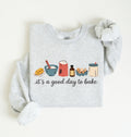 A light heather gray long-sleeve sweatshirt with a graphic of baking ingredients and the text 'it's a good day to bake' printed on the front, styled flat on a light cream background.