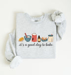 A light heather gray long-sleeve sweatshirt with a graphic of baking ingredients and the text 'it's a good day to bake' printed on the front, styled flat on a light cream background.