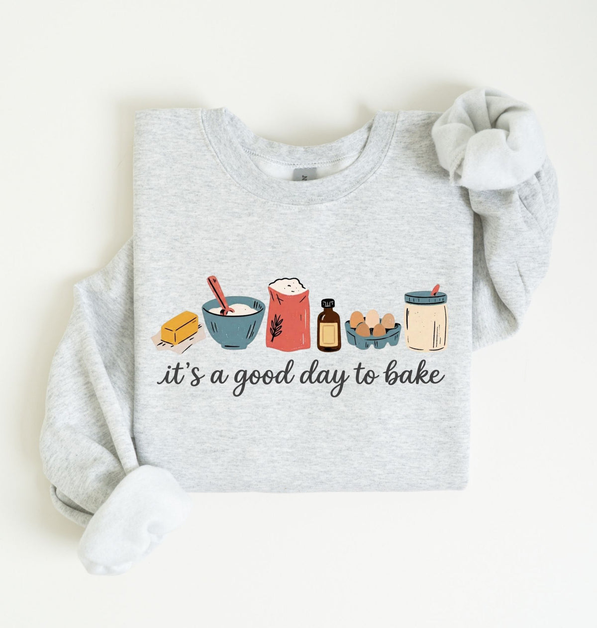 A light heather gray long-sleeve sweatshirt with a graphic of baking ingredients and the text 'it's a good day to bake' printed on the front, styled flat on a light cream background.