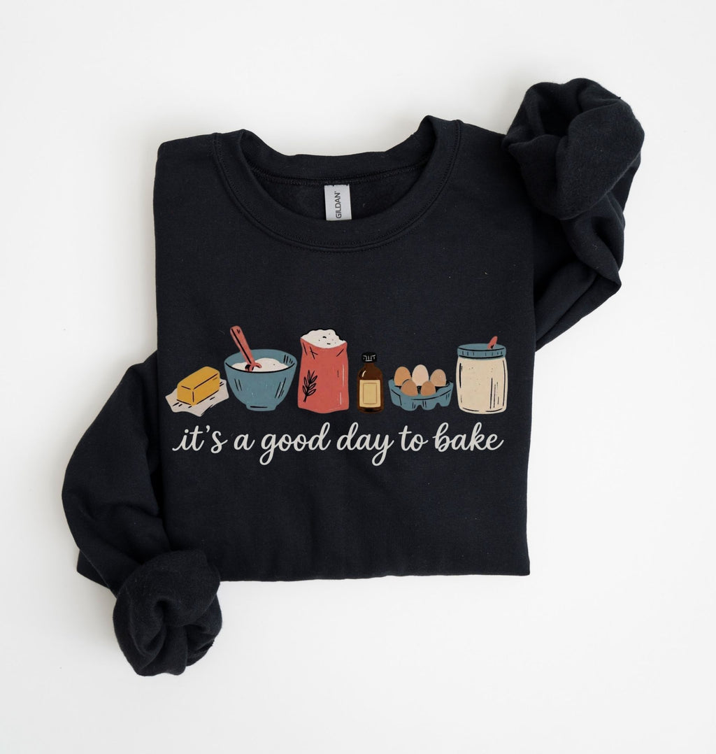 A black long-sleeve sweatshirt with rolled cuffs, featuring a colorful graphic of baking ingredients and the text 'it's a good day to bake' on the front, laid flat on a white background.
