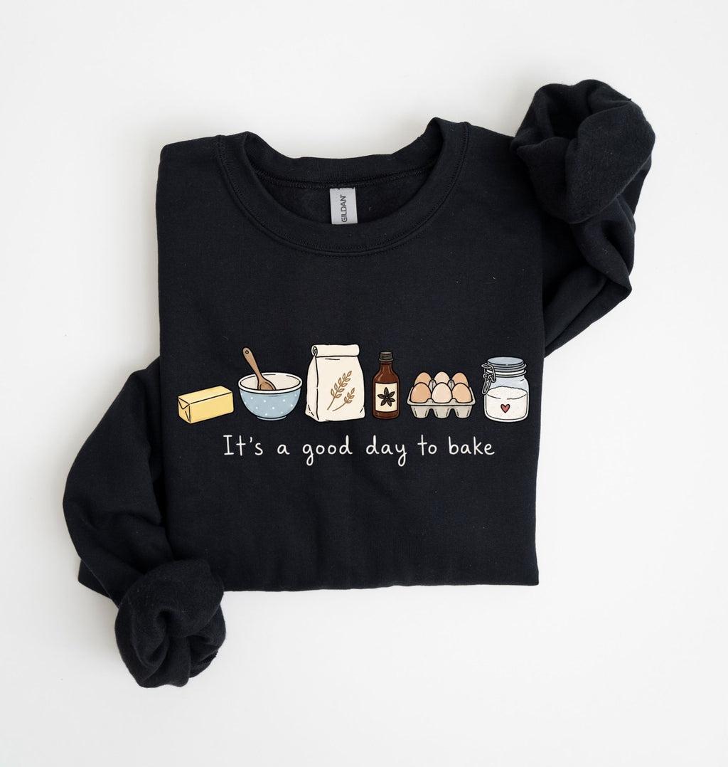 A black long-sleeve sweatshirt with rolled cuffs, featuring a cute baking-themed graphic on the chest with illustrations of a butter stick, bowl, spoon, flour sack, vanilla bottle, eggs, and jar above the text 'It's a good day to bake'.