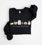 A black long-sleeve sweatshirt with rolled cuffs, featuring a cute baking-themed graphic on the chest with illustrations of a butter stick, bowl, spoon, flour sack, vanilla bottle, eggs, and jar above the text 'It's a good day to bake'.