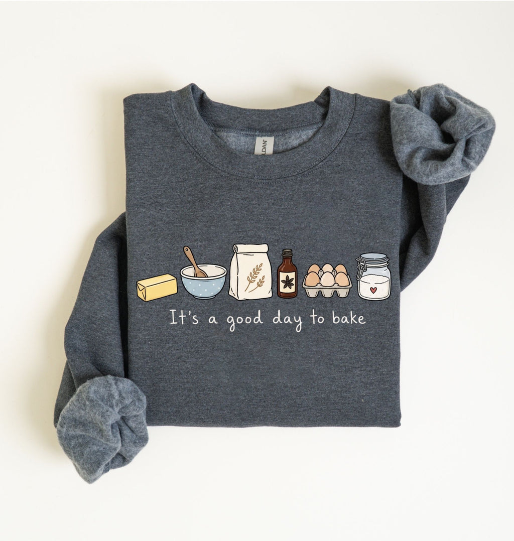 A dark heather gray long-sleeve sweatshirt laid flat on a light cream background. The front has a printed baking-themed graphic with illustrated ingredients and the phrase 'It's a good day to bake' in white script.