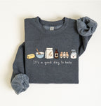 A dark heather gray long-sleeve sweatshirt laid flat on a light cream background. The front has a printed baking-themed graphic with illustrated ingredients and the phrase 'It's a good day to bake' in white script.