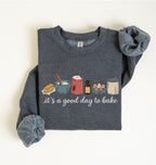 A folded dark heather gray sweatshirt with rolled cuffs, featuring a colorful graphic of baking ingredients and the text 'it's a good day to bake' on the front, set against a plain off-white background.