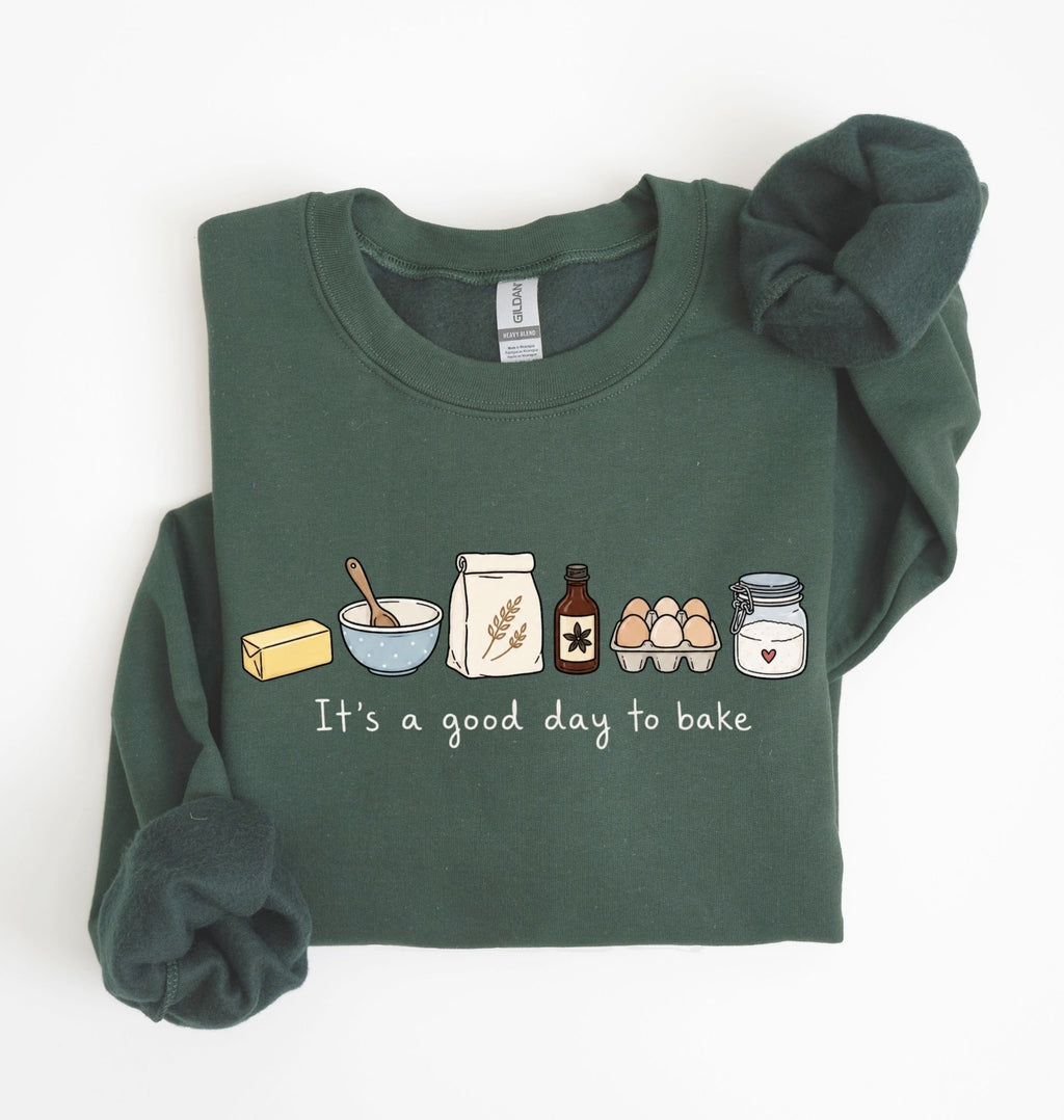 A dark green long-sleeve sweatshirt with a graphic of baking ingredients and the text 'It's a good day to bake' on the front, styled flat on a white background with a matching scrunchie.