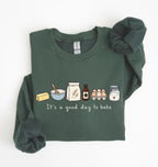 A dark green long-sleeve sweatshirt with a graphic of baking ingredients and the text 'It's a good day to bake' on the front, styled flat on a white background with a matching scrunchie.