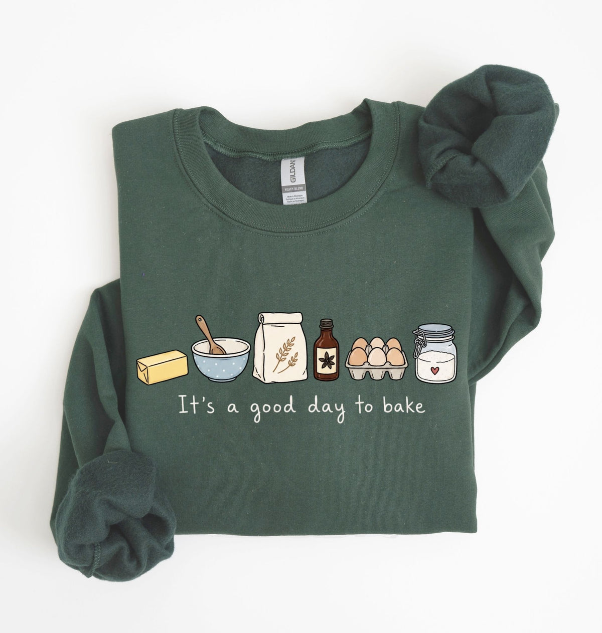 A dark green long-sleeve sweatshirt with a graphic of baking ingredients and the text 'It's a good day to bake' on the front, styled flat on a white background with a matching scrunchie.