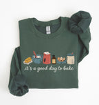A dark green long-sleeve crewneck sweatshirt with a 'it's a good day to bake' graphic on the chest, styled flat on a white background with rolled cuffs and a matching scrunchie.