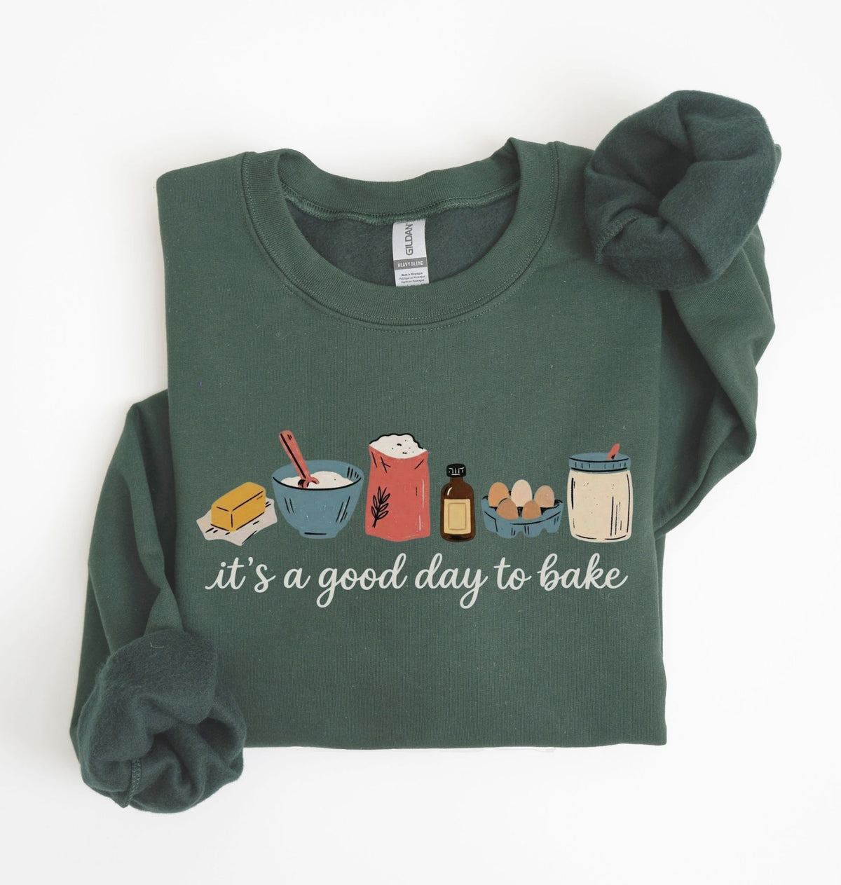 A dark green long-sleeve crewneck sweatshirt with a 'it's a good day to bake' graphic on the chest, styled flat on a white background with rolled cuffs and a matching scrunchie.