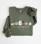 An olive green long-sleeve crewneck sweatshirt with a graphic of baking ingredients and tools printed on the chest, styled with rolled cuffs and a matching fabric scrunchie on a white background.