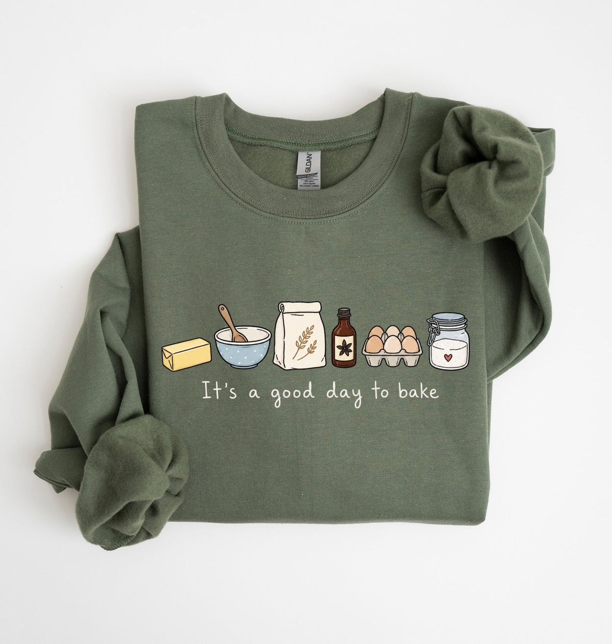 An olive green long-sleeve crewneck sweatshirt with a graphic of baking ingredients and tools printed on the chest, styled with rolled cuffs and a matching fabric scrunchie on a white background.