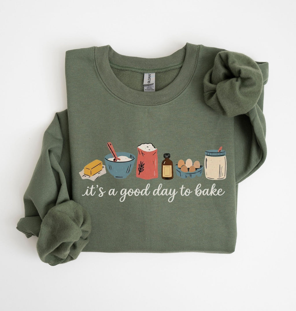 An olive green long-sleeve sweatshirt with rolled cuffs and a matching fabric hair tie, featuring a 'it's a good day to bake' graphic with baking ingredients on a white background.