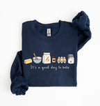 A navy blue long-sleeve sweatshirt with a graphic of baking ingredients and the text 'It's a good day to bake' on the front, displayed flat on a white background.
