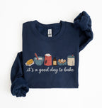 A navy blue long-sleeve crewneck sweatshirt with a 'it's a good day to bake' graphic on the front, featuring baking ingredients like butter, flour, eggs, and a jar of frosting. The sweatshirt is neatly folded on a clean white background.