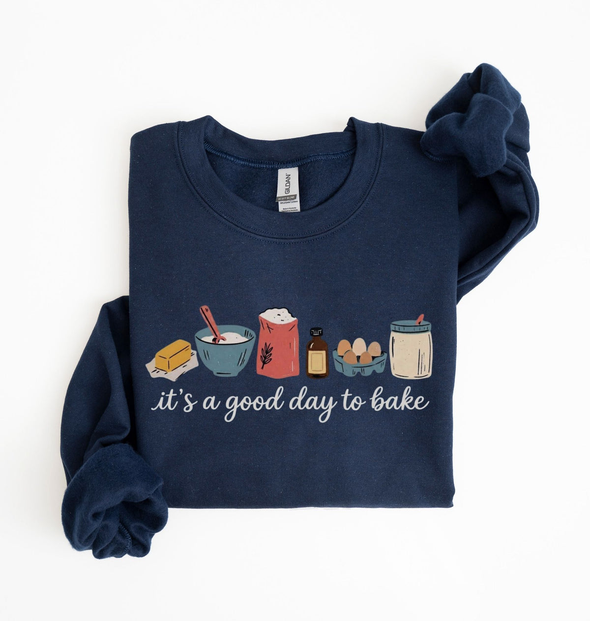 A navy blue long-sleeve crewneck sweatshirt with a 'it's a good day to bake' graphic on the front, featuring baking ingredients like butter, flour, eggs, and a jar of frosting. The sweatshirt is neatly folded on a clean white background.