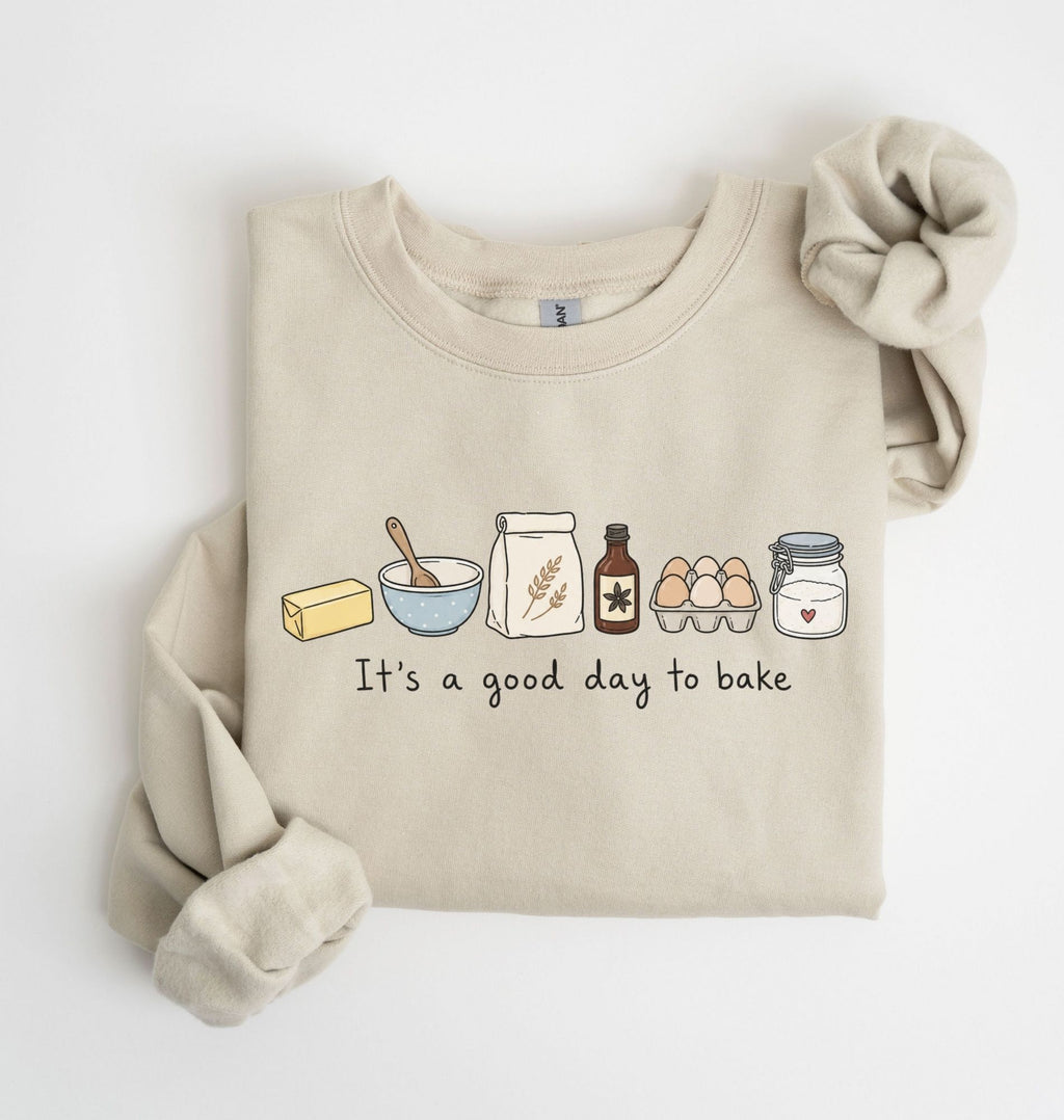 A beige crewneck sweatshirt with a graphic of baking ingredients and the text 'It's a good day to bake', styled flat on a white background with a matching fabric scrunchie.