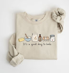 A beige crewneck sweatshirt with a graphic of baking ingredients and the text 'It's a good day to bake', styled flat on a white background with a matching fabric scrunchie.