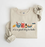 A beige crewneck sweatshirt with a graphic of baking ingredients and the text 'it's a good day to bake' printed on the front, styled flat on a white background.