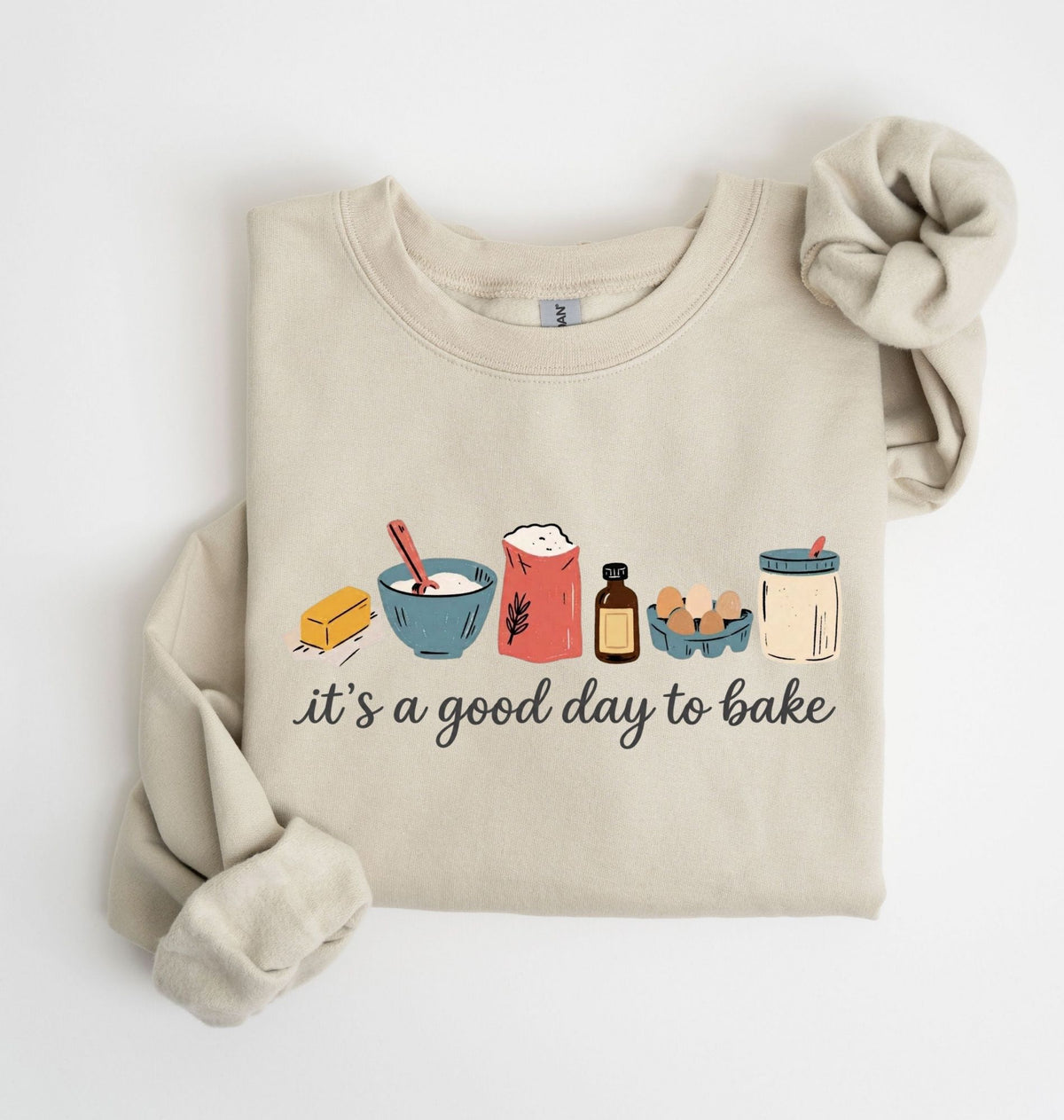 A beige crewneck sweatshirt with a graphic of baking ingredients and the text 'it's a good day to bake' printed on the front, styled flat on a white background.