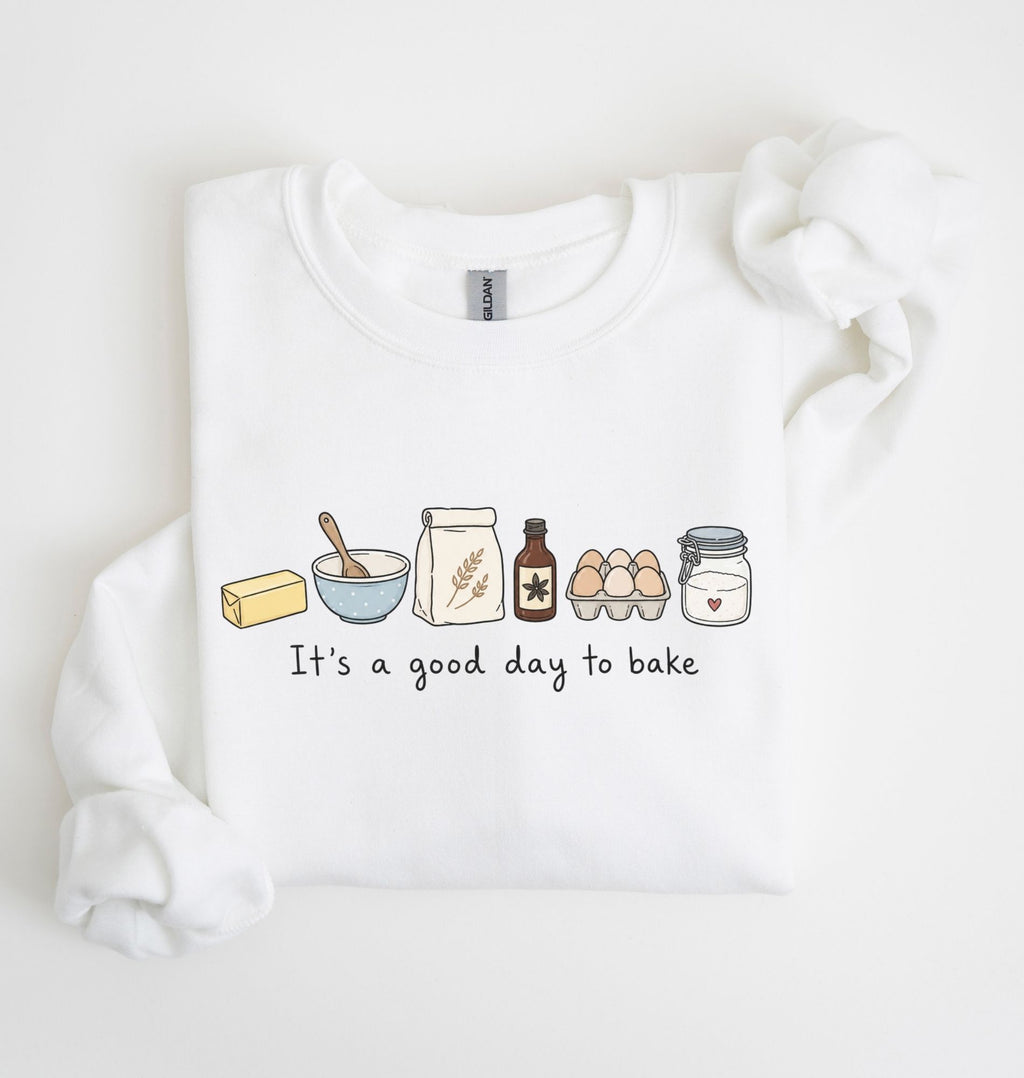 A white long-sleeve sweatshirt with a graphic of baking ingredients and the text 'It's a good day to bake' printed on the front, displayed on a clean white background.