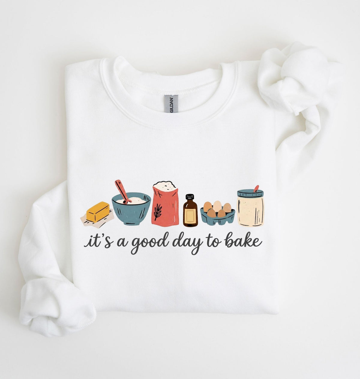 A white long-sleeve crewneck sweatshirt with a graphic print of baking ingredients and the text 'it's a good day to bake' on the front, displayed on a clean white background.