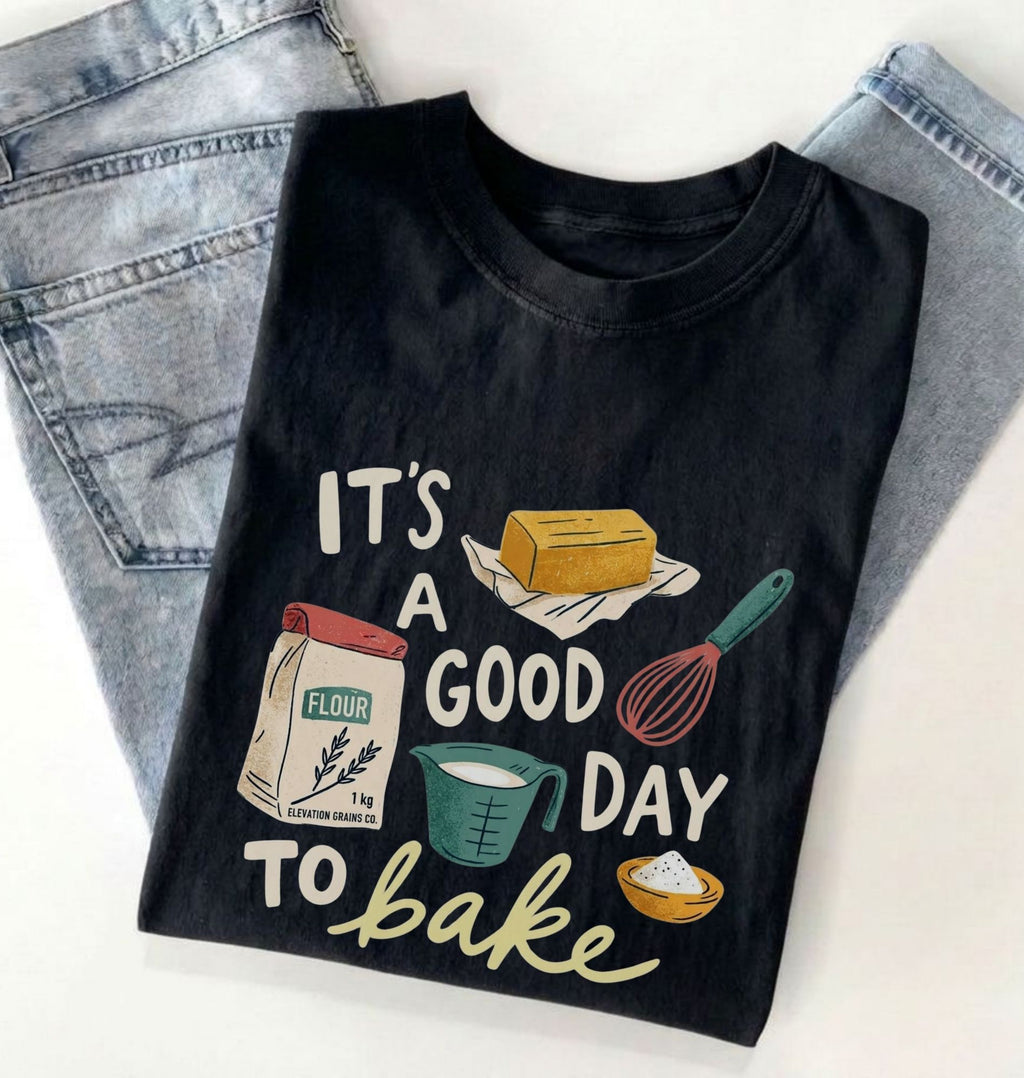 A black graphic t-shirt with a baking-themed print reading 'IT'S A GOOD DAY TO bake' and illustrations of flour, butter, a whisk, and a measuring cup, styled flat-lay on top of light-wash jeans against a white background.
