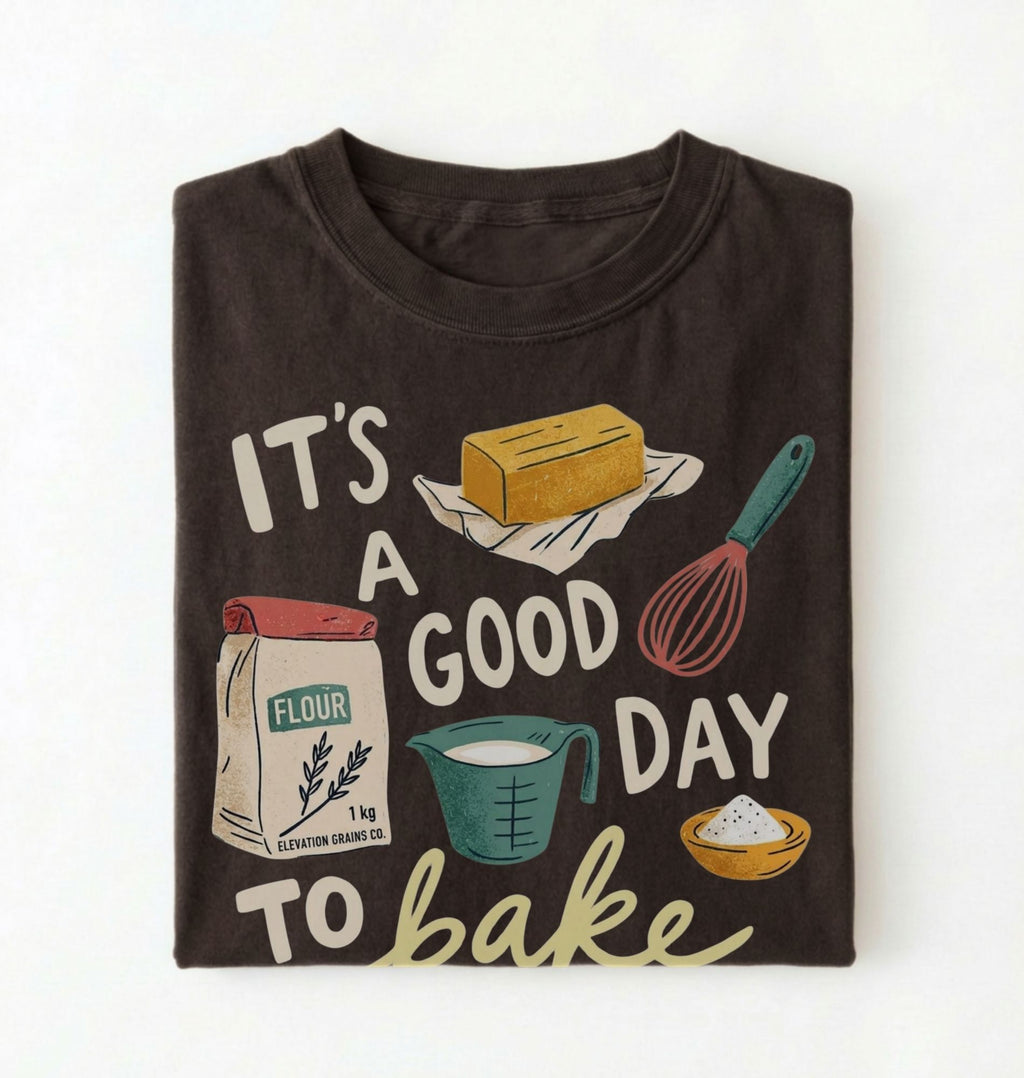 A dark brown folded t-shirt with a graphic print that reads 'IT'S A GOOD DAY TO bake' surrounded by baking illustrations including flour, butter, a whisk, and a measuring cup, shown on a white background.