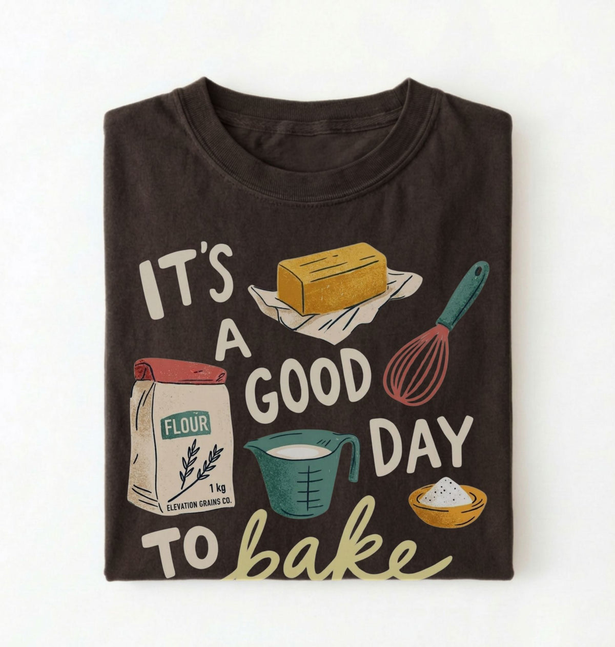 A dark brown folded t-shirt with a graphic print that reads 'IT'S A GOOD DAY TO bake' surrounded by baking illustrations including flour, butter, a whisk, and a measuring cup, shown on a white background.