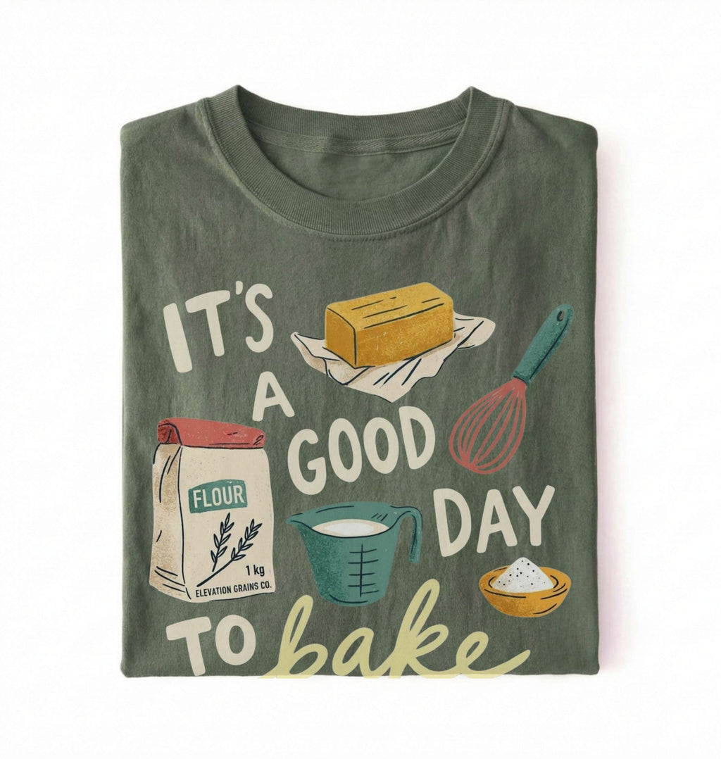 A folded dark olive green t-shirt featuring a colorful graphic print with baking-themed illustrations and the text 'IT'S A GOOD DAY TO bake'.
