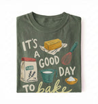 A folded dark olive green t-shirt featuring a colorful graphic print with baking-themed illustrations and the text 'IT'S A GOOD DAY TO bake'.