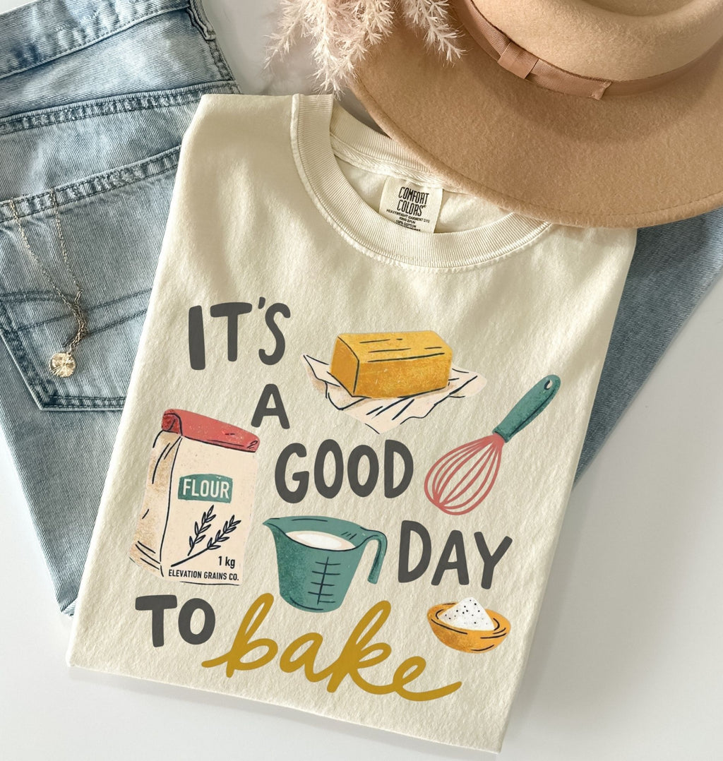 A folded cream-colored graphic T-shirt with a baking-themed print reading 'IT'S A GOOD DAY TO bake' and illustrations of flour, a measuring cup, a whisk, a butter stick, and a bowl, styled on a white surface with blue jeans, a tan hat, and a necklace.