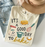 A folded cream-colored graphic T-shirt with a baking-themed print reading 'IT'S A GOOD DAY TO bake' and illustrations of flour, a measuring cup, a whisk, a butter stick, and a bowl, styled on a white surface with blue jeans, a tan hat, and a necklace.