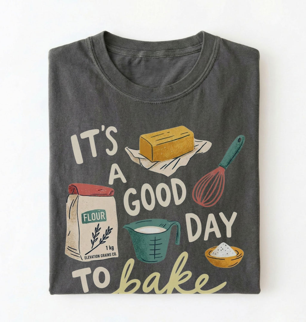 A dark gray folded graphic t-shirt on a white background. The shirt features a retro-style baking illustration with the text 'IT'S A GOOD DAY TO bake' and drawings of a stick of butter, a bag of flour, a measuring cup, a whisk, and a bowl of powder.