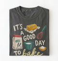 A dark gray folded graphic t-shirt on a white background. The shirt features a retro-style baking illustration with the text 'IT'S A GOOD DAY TO bake' and drawings of a stick of butter, a bag of flour, a measuring cup, a whisk, and a bowl of powder.