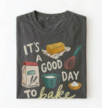 A dark gray folded graphic t-shirt on a white background. The shirt features a retro-style baking illustration with the text 'IT'S A GOOD DAY TO bake' and drawings of a stick of butter, a bag of flour, a measuring cup, a whisk, and a bowl of powder.