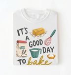 A white short-sleeve t-shirt with a colorful graphic print that reads 'IT'S A GOOD DAY TO bake' accompanied by illustrations of baking ingredients (flour, butter, measuring cup, whisk, bowl). The t-shirt is displayed flat against a clean white background.