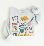 A light heather gray long-sleeve sweatshirt with rolled cuffs, featuring a colorful graphic print that reads 'IT'S A GOOD DAY TO bake' with illustrations of a butter block, flour bag, measuring cup, whisk, and bowl of flour, displayed on a clean off-white background.