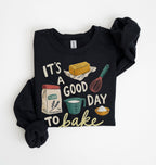 A black long-sleeve crewneck sweatshirt with rolled cuffs, featuring a colorful graphic print that reads 'IT'S A GOOD DAY TO bake' with illustrations of baking ingredients and tools, laid flat on a white background.