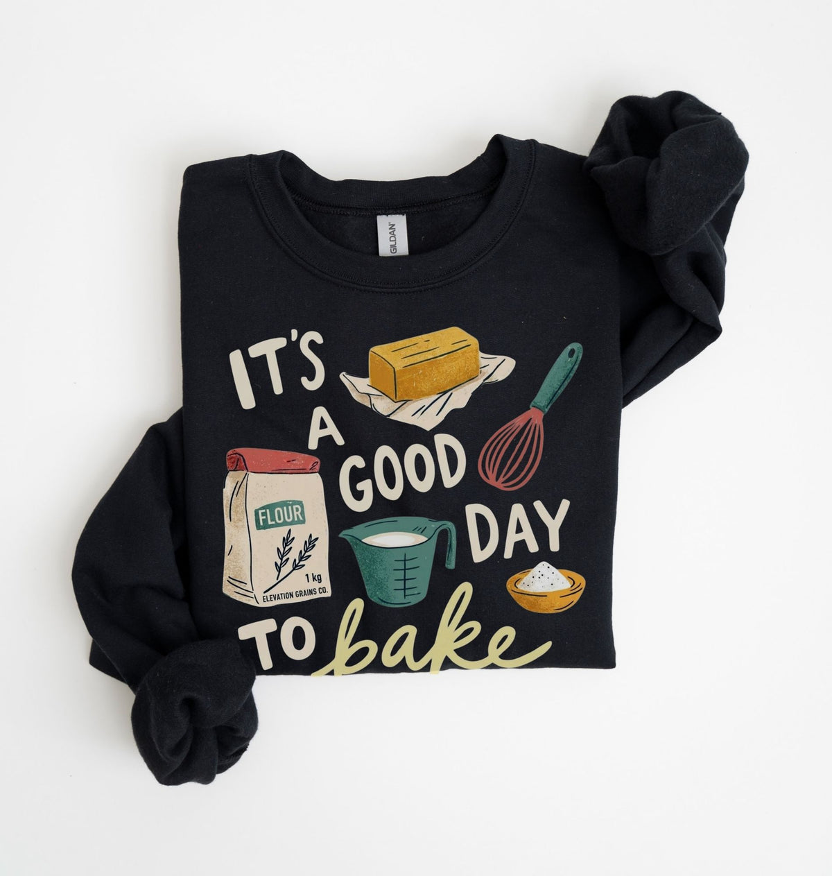 A black long-sleeve crewneck sweatshirt with rolled cuffs, featuring a colorful graphic print that reads 'IT'S A GOOD DAY TO bake' with illustrations of baking ingredients and tools, laid flat on a white background.