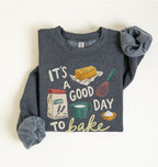 A folded dark heather gray sweatshirt with rolled sleeves, featuring a colorful graphic print that reads 'IT'S A GOOD DAY TO bake' with illustrations of baking ingredients and tools, laid flat on a light cream background.