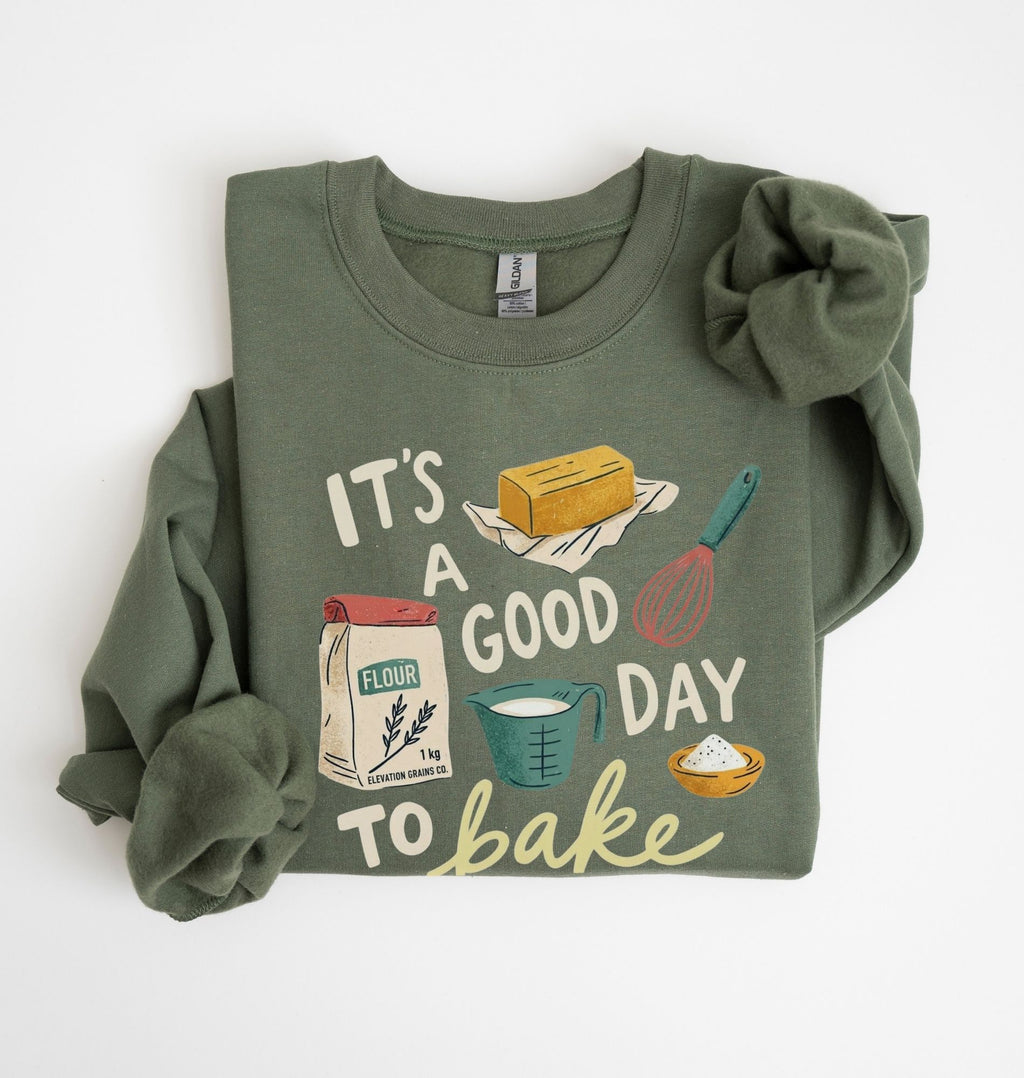 An olive green long-sleeve sweatshirt with a 'It's a Good Day to Bake' graphic print, styled flat on a white background with a matching fabric scrunchie.