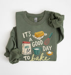 An olive green long-sleeve sweatshirt with a 'It's a Good Day to Bake' graphic print, styled flat on a white background with a matching fabric scrunchie.