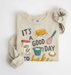 A beige crewneck sweatshirt with rolled sleeves, featuring a colorful graphic print that reads 'IT'S A GOOD DAY TO bake' with illustrations of baking ingredients and tools, displayed on a clean white background.