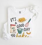 A white crewneck sweatshirt with a colorful graphic print that reads 'IT'S A GOOD DAY TO bake' surrounded by illustrations of baking ingredients (flour, butter, measuring cup, whisk, and pie). The sweatshirt is styled flat on a white background with a matching white fabric scrunchie.