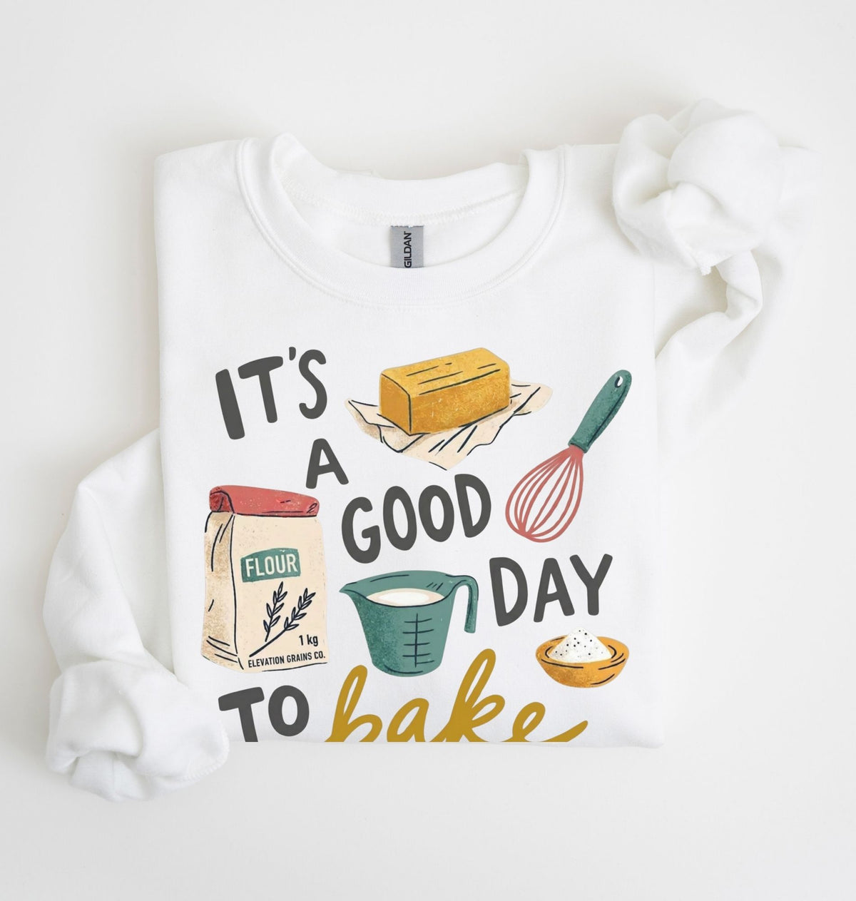 A white crewneck sweatshirt with a colorful graphic print that reads 'IT'S A GOOD DAY TO bake' surrounded by illustrations of baking ingredients (flour, butter, measuring cup, whisk, and pie). The sweatshirt is styled flat on a white background with a matching white fabric scrunchie.