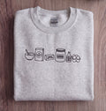 A folded light heather gray sweatshirt on a dark wood background, featuring a black line art graphic of baking ingredients including flour, butter, sugar, syrup, eggs, and a carton.