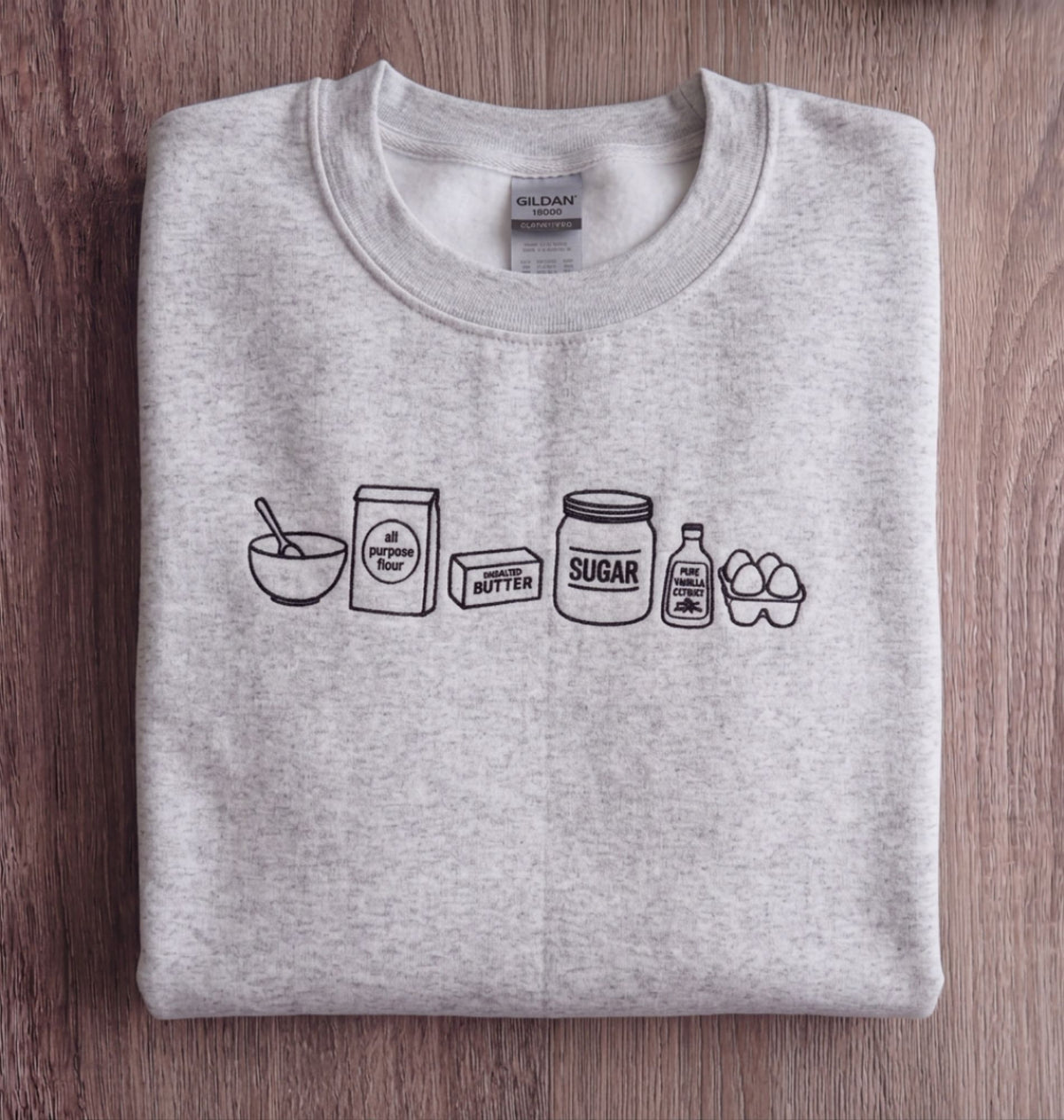 A folded light heather gray sweatshirt on a dark wood background, featuring a black line art graphic of baking ingredients including flour, butter, sugar, syrup, eggs, and a carton.