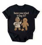 A black short-sleeve graphic t-shirt laid flat on a white background. The shirt features a graphic of a gingerbread man in a white doctor's coat pointing upwards next to a sad gingerbread man, with the text 'have you tried icing it?' printed above them in a beige retro font.